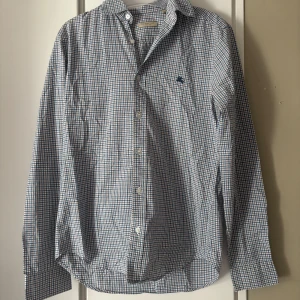 Rutig Burberry skjorta  - Snygg rutig skjorta från Burberry i storlek S. Skjortan är vit, blå och brun med klassiskt rutmönster, knappar framtill och långärmad design. Tillverkad i mjuk bomull och har en broderad Burberry-logga på bröstet. Perfekt för dig som gillar stilrena och tidlösa plagg. Den är lite sliten men inget man tänker på