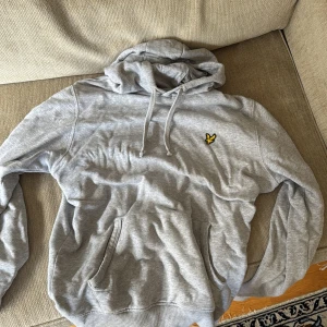 Grå hoodie från Lyle & Scott - Säljer en grå hoodie från Lyle & Scott med klassisk gul fågel-logga på bröstet. Tröjan har huva med snörning, stor magficka och långa ärmar. Perfekt för chill dagar eller när du vill ha en enkel och snygg look.