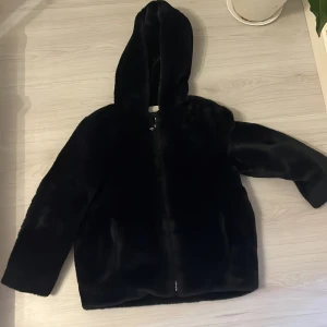 Svart pälsjacka med huva från Zara - Mjuk och fluffig svart pälsjacka från Zara med stor huva och dragkedja framtill. Jackan har en clean look och raka ärmar, perfekt för dig som gillar stilren vibe och vill hålla dig varm. Passar dig som vill ha en cool och mysig jacka med lite edge.