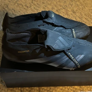 Adidas Predator Elite FT FG fotbollsskor - Säljer ett par svarta Adidas Predator Elite FT FG fotbollsskor med guldiga och silvriga detaljer. Skorna har Hybridtouch 2.0-material, låg profil och snörning som täcks av en elastisk plös. Snygg design med Predator-logga och mönstrad sula
