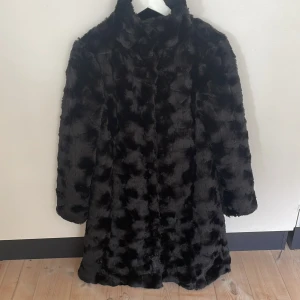 Svart pälsjacka med hög krage - Snygg svart pälsjacka med fluffig känsla och hög krage. Jackan har långärmade och en rak, lite längre passform som ger en cool siluett. Perfekt för dig som vill ha en statement piece i garderoben. Jackan är helsvart och har ett diskret mönster i pälsen.