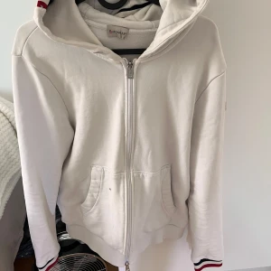 Vit hoodie från Moncler med dragkedja - Säljer en clean vit hoodie från Moncler med hel dragkedja och huva. Tröjan har fickor framtill och snygga ränder i rött, blått och vitt på huvan och ärmsluten. Perfekt för dig som gillar stilrena märkesplagg med sportig vibe.