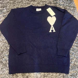 Marinblå stickad tröja från Ami - Snygg marinblå stickad tröja från Ami med stort vitt hjärta och bokstaven A på bröstet. Tröjan har rund hals, ribbade muddar och är långärmad. Perfekt för dig som gillar stilrena detaljer och vill ha något som sticker ut lite extra.