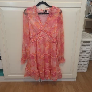 Orange klänning från Vero Moda - Superfin kortklänning från Vero Moda i rosa och orange toner med blommigt mönster. Klänningen har volangdetaljer, v-ringning och långa transparenta ärmar. Materialet är lätt och flowy polyester, perfekt för sommarens alla tillfällen. Söt och färgglad vibe! Orginal pris på hemsidan var 450kr.