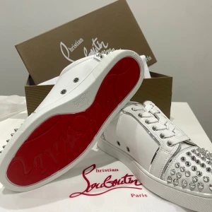 Christian Louboutin vita sneakers med nitar - Säljer ett par sjukt snygga vita sneakers från Christian Louboutin med ikonisk röd sula och silverfärgade nitar på tå och häl. Skorna har snörning, rund tå och är tillverkade i lyxigt skinn. Perfekt för dig som vill sticka ut med en edgy vibe och exklusiv känsla.