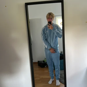 Ljusblå Adidas tracksuit - Säljer en ljusblå Adidas tracksuit med klassiska vita ränder längs ärmarna och broderad logga på bröstet. Aldrig använt. 