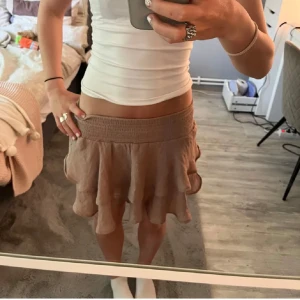 Beige volangkjol med resårmidja - Säljer en beige kortkjol med volanger och bred resår i midjan. Kjolen har en luftig och flowy känsla, perfekt för varma dagar. Materialet känns lätt och skönt mot huden. Enkel att matcha med både toppar och sneakers.