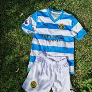 Manchester City Haaland #9 fotbollströja - Manchester City kit med T-shirt och shorts med Haaland och nummer 9 på ryggen. Blå och vita breda ränder, kortärmad med V-ringning. Puma-logga och Etihad Airways tryck på bröstet. Detaljer som 93:20 och motiv i blå ränder. Materialet är lätt polyester.