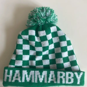 Grönvit Hammarby-mössa för vintern och hösten - Säljer en grön och vit stickad Hammarby-mössa med schackrutigt mönster och stor tofs på toppen. Mössan har uppvikt kant med tydlig Hammarby-text framtill. Perfekt för dig som vill visa ditt stöd på matchen!