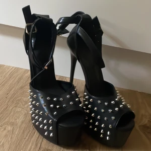 Svarta platåklackar med nitar, strl 41 - Riktigt edgy svarta platåklackar med höga stilettklackar och öppna tår. Skorna har coola silverfärgade nitar över hela ovandelen och breda ankelremmar med spänne. Perfekta för dig som vill sticka ut och älskar statement-skor.