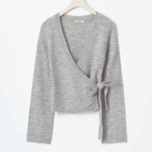 Grå omlottkofta - Säljer en mjuk och mysig grå omlottkofta från Arket. Koftan har en snygg v-ringning, långa ärmar och knyts med en rosett i sidan. Perfekt för lager-på-lager och chill dagar. Passar dig som gillar stilrena och enkla plagg.