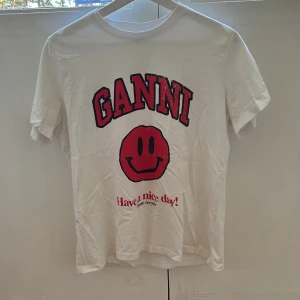 Vit Ganni t-shirt med röd smiley - Vit t-shirt från Ganni. Framsidan har ett stort rött smileytryck och texten 'Have a nice day! Please recycle' i rött och svart. T-shirten är gjord i mjuk bomull och har en relaxed passform, perfekt för en chill look. Den är som ny väldigt sparsamt använd