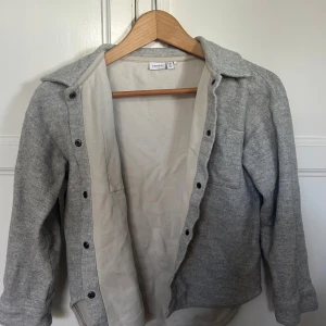 Grå overshirt från Name It - Snygg grå overshirt från Name It med klassisk krage och en bröstficka. Jackan har knappar hela vägen och är tillverkad i mjukt material som känns skönt mot huden. Perfekt för lager-på-lager och chill stil. Passar dig som gillar enkel och clean look.