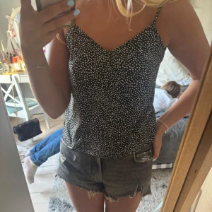 Svart linnetopp med vita hjärtan - Snygg svart linnetopp med tunna axelband och ett mönster av små vita hjärtan. Toppen har en v-ringad hals och ärmlös design som ger en chill och fräsch vibe. Perfekt för varma dagar och enkel att matcha med jeans eller shorts.