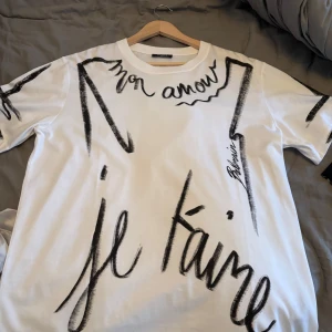 Vit Balmain t-shirt med print - Vit t-shirt från Balmain Paris Couture i bomull med svart handskrivet print på framsidan där det står 'mon amour' och 'je t'aime'. T-shirten har rund hals, korta ärmar och en loose passform. Snygg statement-piece för dig som gillar designer och coola prints. Storleken är som L men den är M egentligen. Och den är äkta men tyvärr inget kvitto 