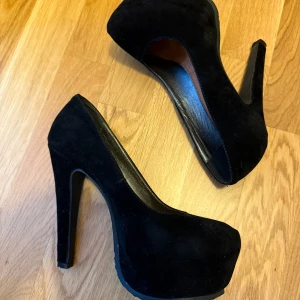 Svarta mocka pumps från Tiamo - Säljer ett par svarta pumps från Tiamo med hög klack och plattform. Skorna har rund tå och är klädda i mjuk mocka som ger en lyxig känsla. Perfekt för dig som vill ha en stilren och klassisk look med extra höjd.