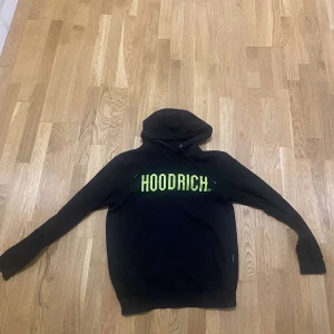 Svart hoodie från Hoodrich - Svart hoodie från Hoodrich med stor neongul logga på bröstet. Tröjan har huva med snörning och långa ärmar. Perfekt för dig som gillar streetwear och vill sticka ut med en fet logga.