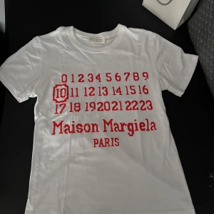 Maison Margiela vit t-shirt med tryck - Maison Margiela t-shirt i vit bomull med rött grafiskt tryck framtill. Trycket består av siffror och Maison Margiela Paris-logga. Klassisk rund hals och korta ärmar. Perfekt för dig som gillar statement-plagg och designerstil.