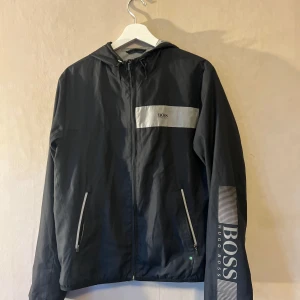 Svart vindjacka från Hugo Boss - Svart vindjacka från Hugo Boss med huva och dragkedja framtill. Jackan har gråa detaljer på bröstet och ärmarna, samt tydlig BOSS-logga på vänster ärm. Två fickor med dragkedja och regular fit. Perfekt för blåsiga dagar och snygg streetstil.