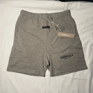 Grå Essentials shorts - Gråa shorts från Essentials Fear of God med elastisk midja och snörning. Mjukt bomullsmaterial, enkel design med logga på vänster ben och bakficka. Perfekta för chill dagar eller streetwear-stil.