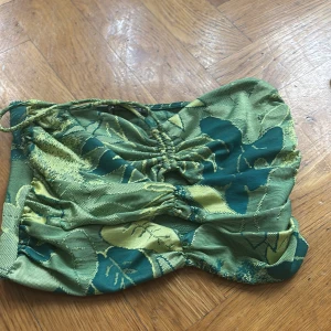 Grön bandeau topp från Zara - Säljer en grön bandeau topp från Zara med blommigt mönster i olika gröna och gula nyanser. Toppen har snörning framtill och är axelbandslös med en croppad passform. Materialet är tjockt och har en lätt glansig yta som känns lyxig.