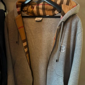 Grå Burberry hoodie med dragkedja - Säljer en grå hoodie från Burberry med klassiskt rutigt foder i huvan och insidan av dragkedjan. Tröjan har en stor B-logga på bröstet, snörning i huvan och praktiska fickor. Nog den skönaste hoddien jag ägt men börjar bli för liten. Den är i storlek S men passar även medium. Skicket är bra och kvitto finns. Hör av er vid funderingar!