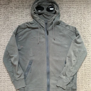 Grå hoodie med goggles från C.P. Company - Säljer en grå hoodie från C.P. Company med ikoniska goggles i huvan. Tröjan har dragkedja framtill, två fickor med zip och tumhål i mudden. Perfekt för dig som vill sticka ut med streetwear-vibe och coola detaljer. Huvan har snörning och goggles med logga. OBS ingen frakt, säljer endast i Göteborgsområdet men kan också bytas mot något annat Stone Island eller C.P. Company 