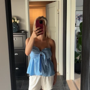Blå axelbandslös topp med knyt - Säljer en ljusblå axelbandslös topp med volangkant och snygg knytning framtill. Toppen har en luftig och flowy passform som ger en chill vibe. Perfekt för varma dagar och enkel att styla med shorts eller jeans.