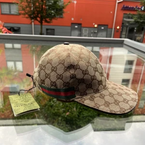 Gucci beige keps med GG-mönster - Snygg beige keps från Gucci med klassiskt GG-monogram över hela kepsen och en grön-röd rand på sidan. Justerbar rem baktill i svart läder. Materialet är slitstark bomull och kepsen har en böjd skärm. Perfekt för dig som gillar exklusiva accessoarer.
