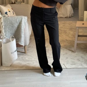 Svarta randiga kostymbyxor Madlady - Snygga svarta kostymbyxor från Madlady med smala vita ränder. Byxorna har rak passform, hög midja och stängs med dragkedja, knapp och hyska. Perfekta för en clean och stilren look. Materialet känns lätt och bekvämt.