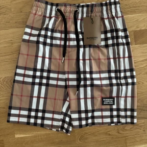 Rutig Burberry shorts med snörning - Snygga shorts från Burberry med klassiskt rutigt mönster i beige, svart, vitt och rött. Modellen har elastisk midja med svart snörning och Burberry-logga framtill. Perfekta för sommaren och har en loose passform som ger skön vibe.