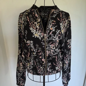 Svart blommig bomberjacka Cubus XS - Snygg svart bomberjacka från Cubus i storlek XS med blommigt mönster i rosa, vitt och gult. Jackan har dragkedja framtill, ribbad krage och muddar. Tillverkad i mjuk polyester som känns lätt och skön. Perfekt för dig som gillar en chill och trendig stil.