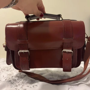 Brun axelväska i skinn med spännen - Säljer en snygg brun axelväska i skinn med klassiska spännen i silverfärg. Väskan har ett handtag upptill, justerbar axelrem och flera fack, inklusive ett mindre ytterfack med spänne. Perfekt för dig som vill ha en stilren och praktisk accessoar.