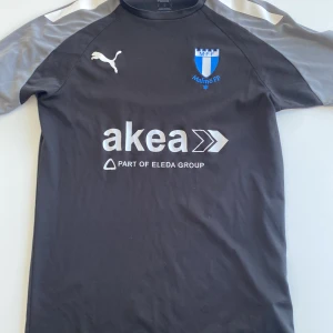 Malmö FF träningströja Puma - Säljer en svart och grå Malmö FF träningströja från Puma med korta ärmar. Tröjan har klubbmärke och sponsortryck på bröstet samt Puma-logga. Perfekt för fotbollsträning eller supporterliv.