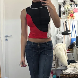 Färgglad croppad polotopp - Säljer en unik croppad polotopp med höghalsad krage. Toppen har blockmönster i svart, rött, vitt och beige med kontrastsömmar. Ärmlös och figurnära passform, perfekt för att sticka ut. Står M/L på lappen men passar som XS/S