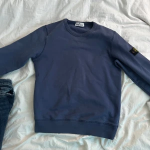 Blå sweatshirt från Stone Island Junior - Säljer en blå sweatshirt från Stone Island Junior med klassisk rund halsringning och ikonisk logotyp-patch på ärmen. Tröjan har en enkel och stilren design med ribbade muddar vid ärmslut och nederkant. Pris relativit fast, skulle kunna diskuteras.