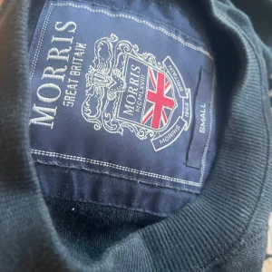 Mörkblå sweatshirt från Morris - Mörkblå sweatshirt från Morris med broderad logga och brittisk flagga på bröstet. Tröjan har rund halsringning och långa ärmar. Perfekt för en stilren och klassisk look.