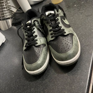 Nike Dunk Low i svart och grön - Säljer ett par Nike Dunk Low sneakers i svart och grön med vit sula. Skorna har klassisk snörning, svart swoosh och perforerad tå. Perfekta för dig som gillar streetwear och vill ha en stilren sneaker med cool färgkombo.