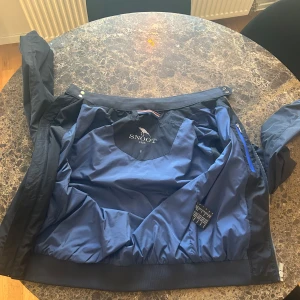 Marinblå vindjacka från Snoot - Säljer en marinblå vindjacka från Snoot med dragkedja och tryckknappar. Jackan har flera fickor och detaljer i silverfärg. Perfekt för blåsiga dagar och har en klassisk, sportig look. Har knappt använt den.