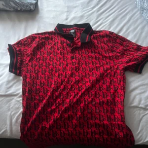 Röd och svart mönstrad t-shirt från Black Squad - Säljer en röd t-shirt från Black Squad med svart mönster över hela tröjan. T-shirten har svart krage och svarta muddar vid ärmarna. Slim fit-modell som passar perfekt till en trendig stil.