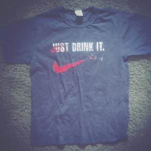 Mörkblå t-shirt med tryck 'Just Drink It' - Mörkblå t-shirt med vitt och rött tryck på bröstet där det står 'Just Drink It' och en röd vinflaska som liknar en Nike-logga. Klassisk passform och rund halsringning. Perfekt för dig som gillar humoristiska plagg.