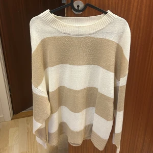 Beige och vit randig stickad tröja - Säljer en oversized stickad tröja med breda beige och vita ränder. Tröjan har lång ärm och en bred ribbad krage. Perfekt för en avslappnad och trendig look.