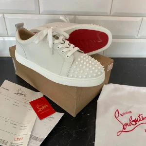 Christian Louboutin - Säljer ett par vita sneakers från Christian Louboutin med ikonisk röd sula och vita nitar på tån. Skorna har snörning och är tillverkade i skinn. Perfekta för dig som vill sticka ut med en lyxig och edgy stil.