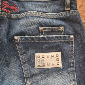 Philipp Plein Jeans - slim fit - w29 - Säljer ett par blå jeansbyxor från Philipp Plein med slitningar och snygga detaljer. Byxorna har knappar i gylfen, röda broderier och en metallplatta med nitar på bakfickan. Klassisk femficksmodell med raka ben.