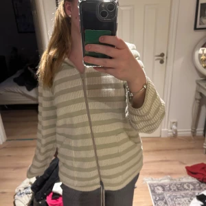 Beige och vit randig kofta med dragkedja - Mysig kofta i beige och vitt med horisontella ränder och dragkedja framtill. Tröjan har långa ärmar och en avslappnad passform, perfekt för lager-på-lager. Snygg att matcha med jeans eller mjukisbyxor.