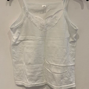 Vit linnetopp med spetsdetalj - Säljer en vit linnetopp med smala axelband och vacker spetsdetalj framtill. Toppen är ärmlös och har en enkel, stilren design som passar perfekt till sommaren.