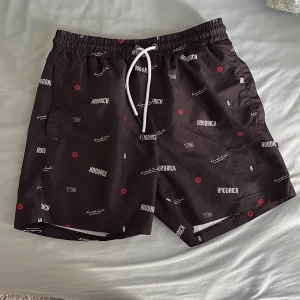 Svarta badshorts från Hoodrich med tryckt logga - Svarta shorts från Hoodrich med vitt och rött tryck över hela tyget. Elastisk midja med vit snörning och innerfoder i mesh. Perfekta för sommaren och har en modern streetwear-look.