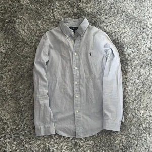 Ljusblå skjorta från Ralph Lauren - Säljer en klassisk ljusblå skjorta från Ralph Lauren med button-down krage och broderad logga på bröstet. Skjortan har långa ärmar och vita knappar framtill. Perfekt för en stilren look.