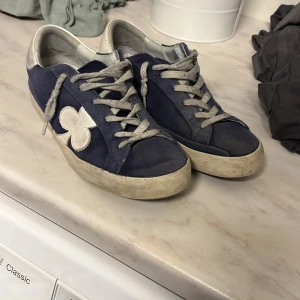 Blå sneakers från The Gallerist - Säljer ett par blå sneakers från The Gallerist med vita detaljer och snörning. Skorna har en vit sula och en stor vit klöverdetalj på sidan. Perfekta för dig som gillar en sportig och avslappnad stil box ingår och ska borsta upp mockan och tvätta dom 