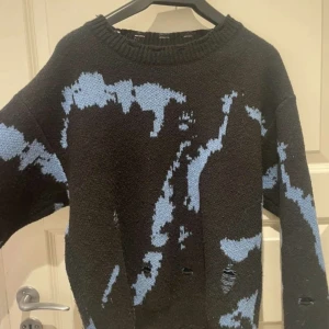 Svart stickad tröja med blått mönster - Cool svart stickad tröja med abstrakt blått mönster. Tröjan har en avslappnad passform, rund halsringning och långa ärmar. Perfekt för dig som gillar en unik och edgy stil.